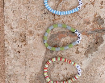 Puede incluir: Cinco pulseras de cuentas de colores con nombres escritos con cuentas de letras blancas. Las pulseras son en tonos marrón, azul, verde, rojo y naranja. Los nombres en las pulseras son John, Sarah, Pope, Kie y JJ.