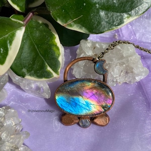 May include: A copper-toned pendant necklace featuring an oval labradorite stone with iridescent blue, purple, and yellow hues. The pendant has a moon charm and a small, round stone at the base. The necklace is displayed on a lavender surface.