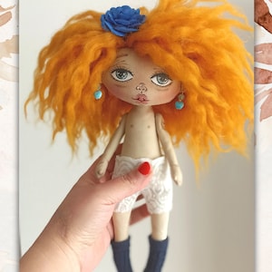 May include: A handmade doll with orange curly hair, a blue flower, turquoise earrings, and white lace shorts. The doll is wearing blue socks and is being held by a hand.