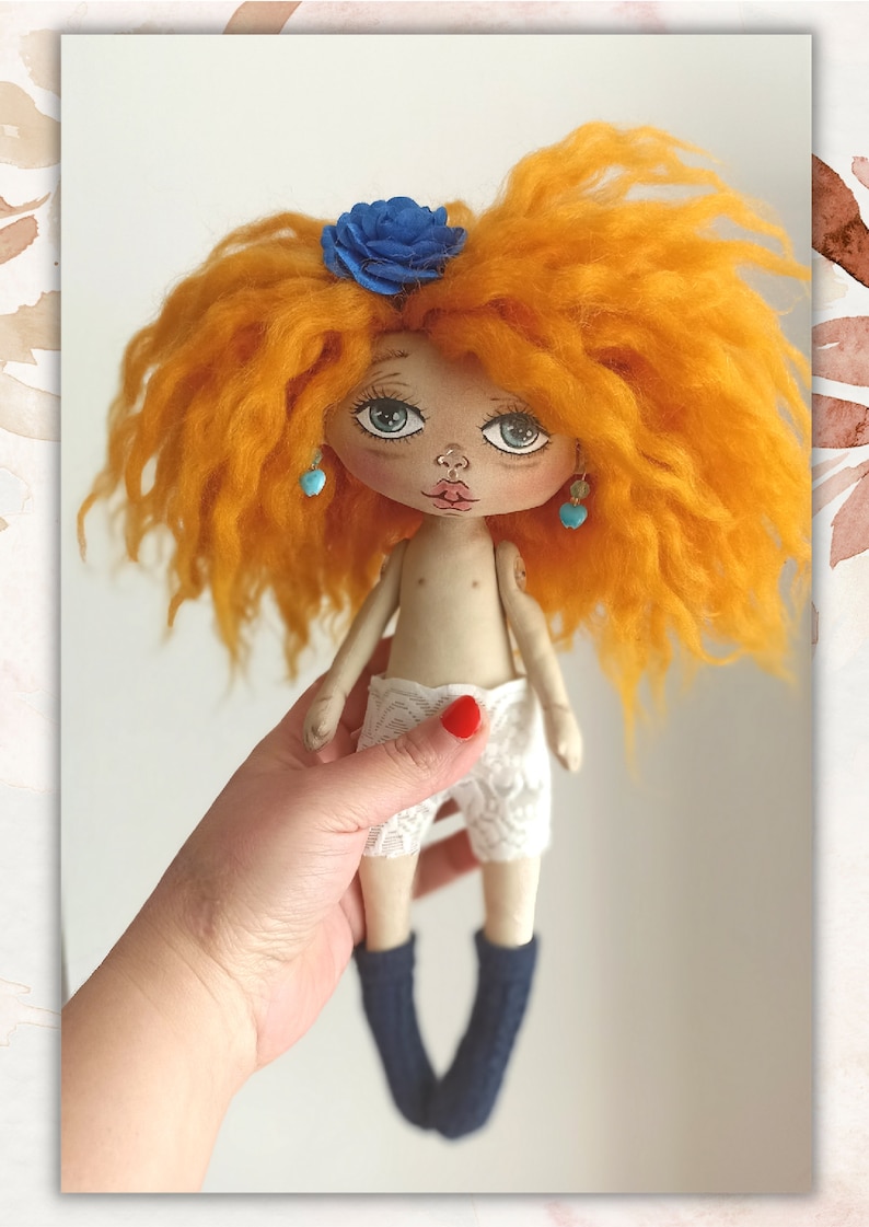 May include: A handmade doll with orange curly hair, a blue flower, turquoise earrings, and white lace shorts. The doll is wearing blue socks and is being held by a hand.
