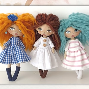 May include: Three handmade rag dolls with different coloured hair and clothing. The doll on the left has orange hair, a blue and white gingham dress, and blue socks. The doll in the middle has brown hair, a white dress with brown buttons, and brown socks. The doll on the right has blue hair, a pink and white striped dress, and white socks.