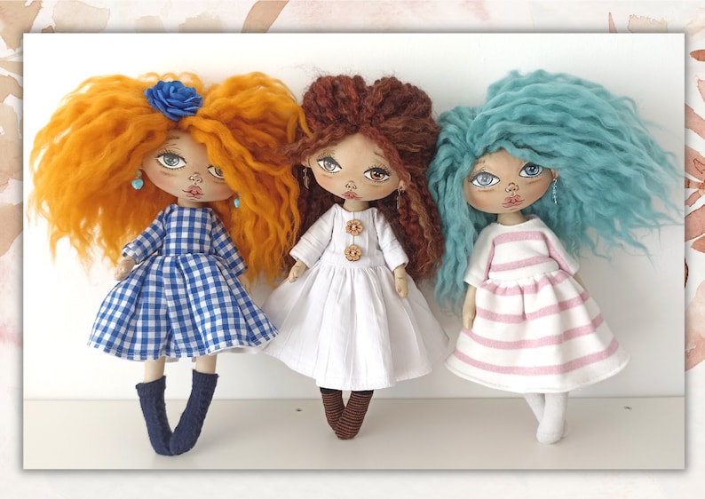 May include: Three handmade rag dolls with different coloured hair and clothing. The doll on the left has orange hair, a blue and white gingham dress, and blue socks. The doll in the middle has brown hair, a white dress with brown buttons, and brown socks. The doll on the right has blue hair, a pink and white striped dress, and white socks.