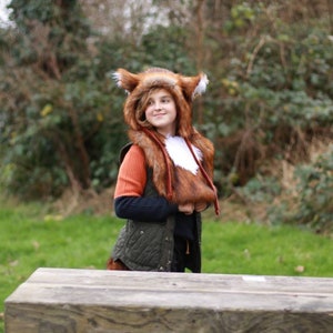 May include: A young person wearing a brown and white fox costume with a green quilted vest. The person is standing in front of a wooden table with a green grassy background.