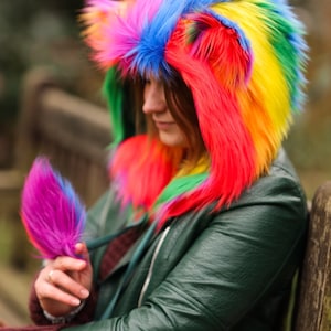 Peut inclure: Une personne portant une veste à capuche en fausse fourrure arc-en-ciel avec un boa en plumes violet dans la main.