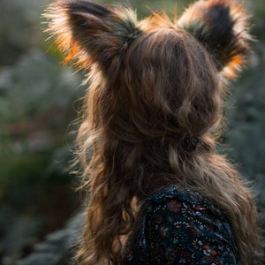 May include: A person wearing a set of faux fox ears with brown and black fur. The person is wearing a dark floral print dress. The image is taken outdoors with soft, natural lighting.