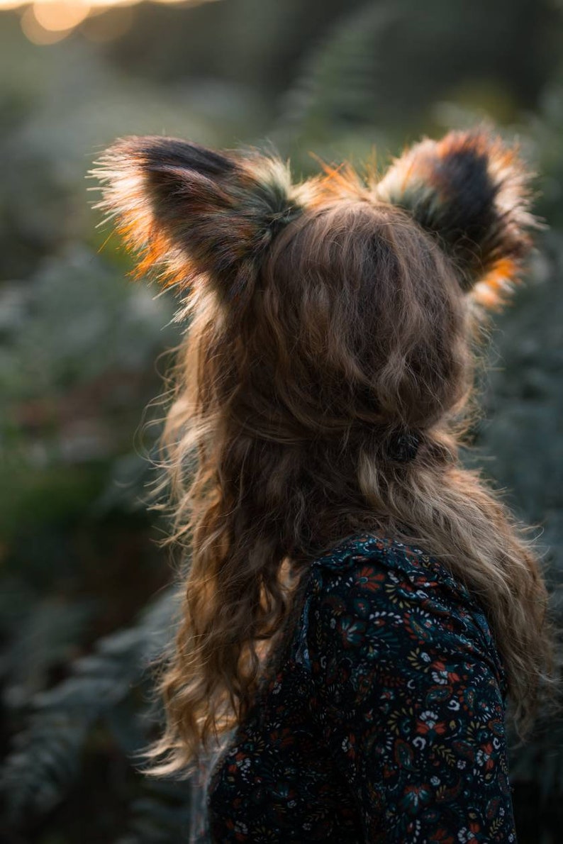May include: A person wearing a set of faux fox ears with brown and black fur. The person is wearing a dark floral print dress. The image is taken outdoors with soft, natural lighting.
