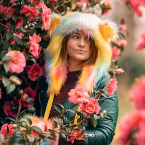 Peut inclure: Une femme portant un chapeau à capuche en fausse fourrure colorée avec une veste en cuir vert, debout devant un buisson de fleurs de camélia roses.