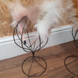 May include: Faux fur cat ears in pink and white, attached to a thin headband. The ears are displayed on a decorative wire head stand against a light wood background.