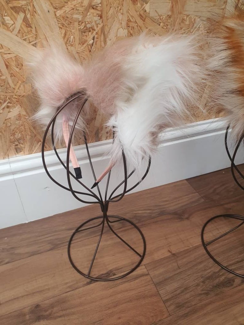 May include: Faux fur cat ears in pink and white, attached to a thin headband. The ears are displayed on a decorative wire head stand against a light wood background.