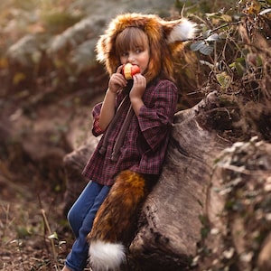 May include: A child wearing a fox costume, including a furry hood, tail, and plaid shirt, is eating an apple. The costume has a brown and white color scheme. The child is sitting on a log in a forest setting.