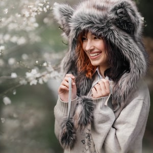 Puede incluir: Un sombrero de piel sint&eacute;tica con orejeras y una bufanda a juego. El sombrero es gris y marr&oacute;n, con una textura esponjosa. La persona lleva un su&eacute;ter gris claro.