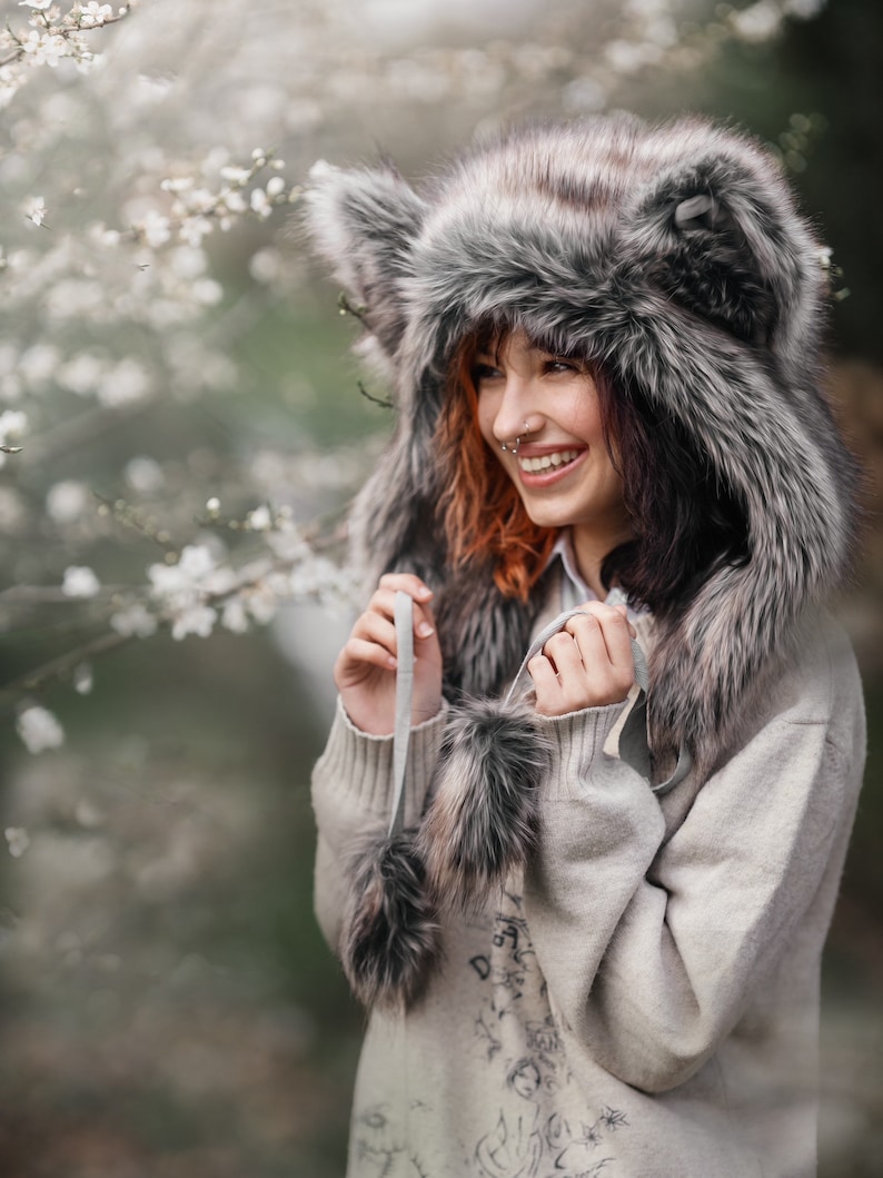 Puede incluir: Un sombrero de piel sint&eacute;tica con orejeras y una bufanda a juego. El sombrero es gris y marr&oacute;n, con una textura esponjosa. La persona lleva un su&eacute;ter gris claro.
