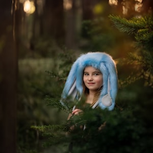 Peut inclure: Une jeune personne portant un chapeau de lapin bleu avec de longues oreilles en fourrure. Le chapeau est doublé de blanc et chaque oreille est surmontée d'un pompon blanc. La personne est debout dans un environnement forestier, entourée d'arbres et de feuillage verts.
