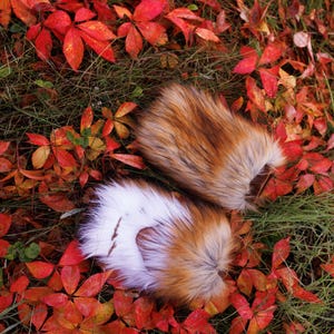 May include: Two fluffy fox tail keychains with white tips, lying on a bed of red and orange autumn leaves.
