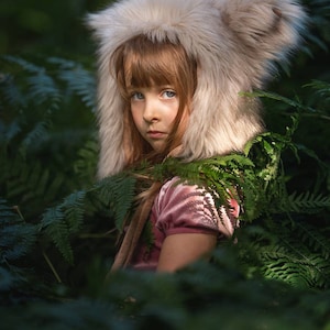 May include: A young girl wearing a furry, light brown hooded hat with large ears, looking directly at the camera. She is wearing a pink velvet dress and is surrounded by green ferns.