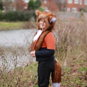 May include: A person wearing a brown and white fox costume with a long tail. The costume includes a hooded hat with furry ears and a matching scarf. The person is standing in front of a body of water and some trees.