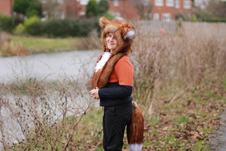 May include: A person wearing a brown and white fox costume with a long tail. The costume includes a hooded hat with furry ears and a matching scarf. The person is standing in front of a body of water and some trees.