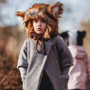 May include: A young child wearing a brown and orange faux fur fox hat and a gray coat with a brown scarf. The child is looking at the camera with a serious expression.