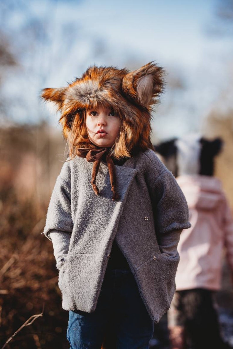 May include: A young child wearing a brown and orange faux fur fox hat and a gray coat with a brown scarf. The child is looking at the camera with a serious expression.