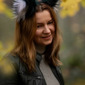 May include: A woman wearing a headband with faux fur fox ears in shades of green, white, and black. She is wearing a black leather jacket and a grey sweater. The background is blurred with autumn colours.
