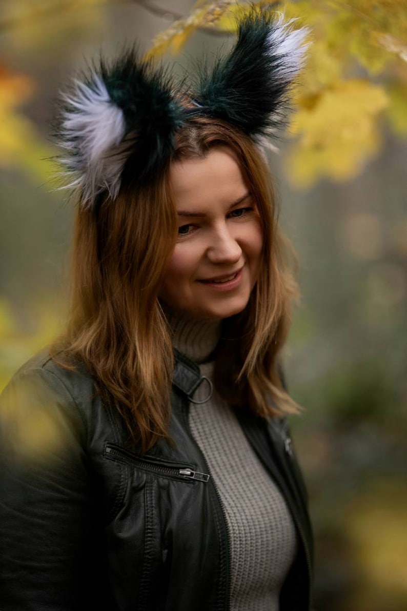 May include: A woman wearing a headband with faux fur fox ears in shades of green, white, and black. She is wearing a black leather jacket and a grey sweater. The background is blurred with autumn colours.