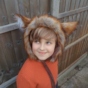 May include: A child wearing a brown and orange faux fur fox hat with large ears. The hat has a brown velvet tie. The child is wearing an orange sweater.