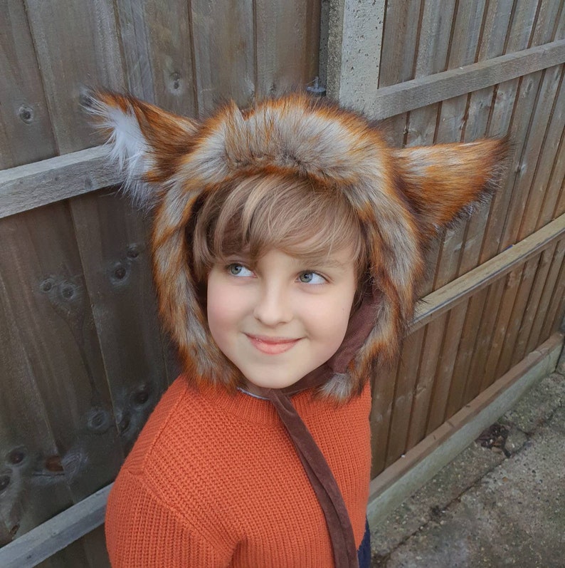 May include: A child wearing a brown and orange faux fur fox hat with large ears. The hat has a brown velvet tie. The child is wearing an orange sweater.