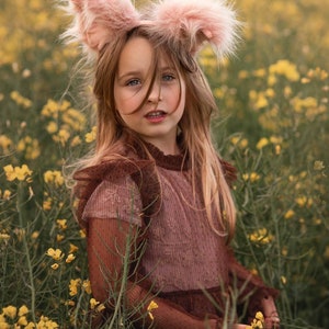 May include: A young person wearing pink bunny ears and a brown dress stands in a field of yellow flowers. The dress has a ribbed texture and ruffled sleeves. The bunny ears are fluffy and light pink.