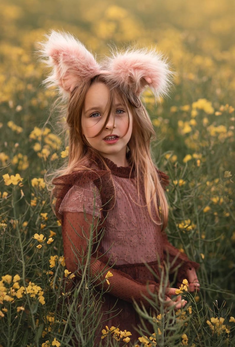 May include: A young person wearing pink bunny ears and a brown dress stands in a field of yellow flowers. The dress has a ribbed texture and ruffled sleeves. The bunny ears are fluffy and light pink.