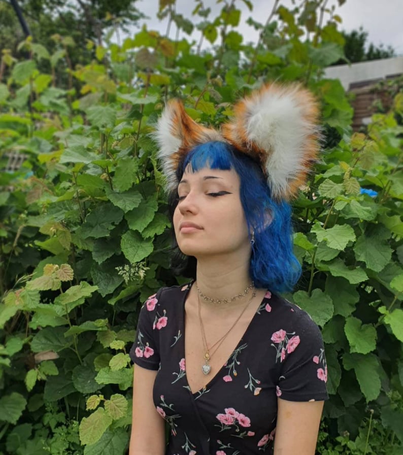 May include: A headband with fox ears made of orange and white faux fur. The person in the image is wearing a black dress with pink floral patterns and has blue hair. The ears are a striking accessory.
