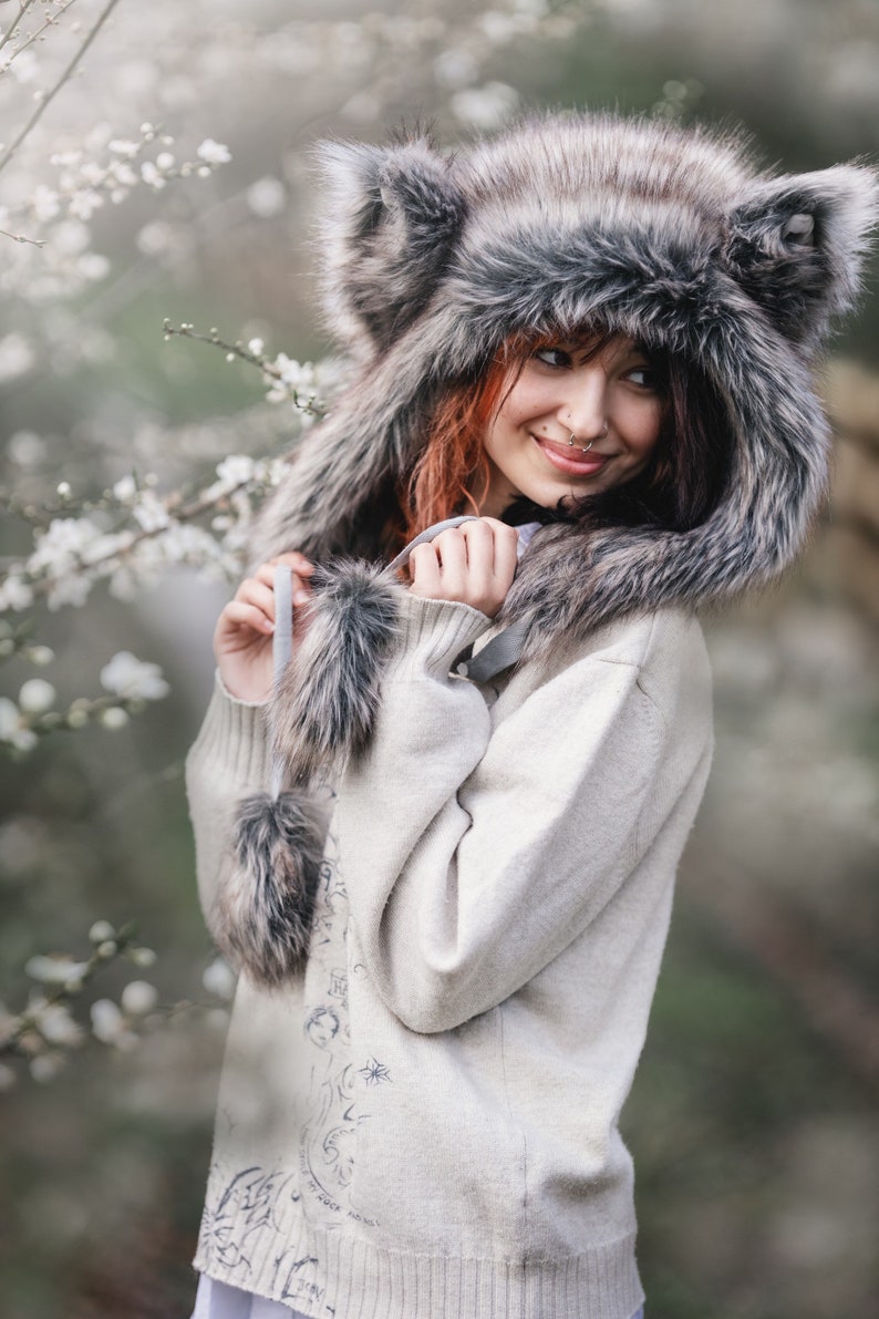 Puede incluir: Un gorro de piel sint&eacute;tica con orejas grandes y erguidas y un ribete de piel a juego. El gorro es gris y marr&oacute;n, y se lleva con un su&eacute;ter de color claro. El su&eacute;ter tiene un dise&ntilde;o decorativo en la espalda y est&aacute; adornado con pompones de piel.