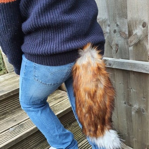 May include: A person wearing blue jeans and a dark blue sweater with a long, fluffy, red and white fox tail attached to the back of their waist.