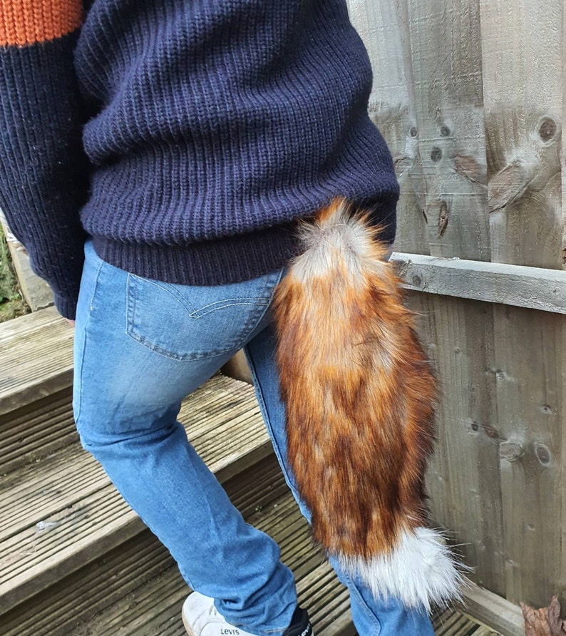 May include: A person wearing blue jeans and a dark blue sweater with a long, fluffy, red and white fox tail attached to the back of their waist.