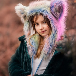Peut inclure: Une jeune personne portant une veste en fourrure verte avec un chapeau en fausse fourrure arc-en-ciel et rose avec de grandes oreilles. Le chapeau est orné d'un ruban bleu.