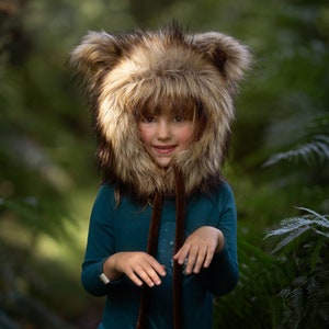 May include: A child wearing a brown and tan faux fur hooded hat with large ears. The hat is pulled down over the child's head, covering their forehead and eyebrows. The child is wearing a teal blue long-sleeved shirt and is smiling.