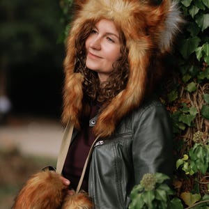 May include: A faux fur hood featuring pointed fox-like ears in shades of orange, brown, and white, paired with matching faux fur hand warmers attached by a beige strap, worn over a dark green jacket and a dark maroon shirt.