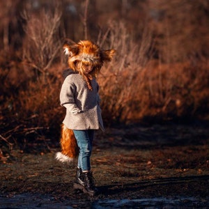 May include: A young person wearing a brown and white fox costume with a furry tail. The person is standing in a field with brown grass and trees in the background.