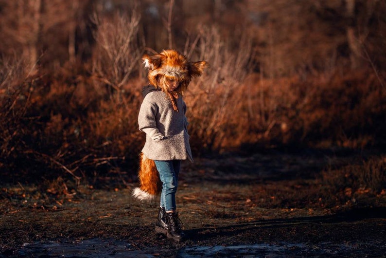 May include: A young person wearing a brown and white fox costume with a furry tail. The person is standing in a field with brown grass and trees in the background.