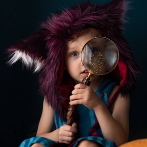 May include: A child wearing a burgundy faux fur hood with pointed ears and a blue striped outfit. The child is holding a magnifying glass with a brass handle, looking through it. The background is a dark teal color.