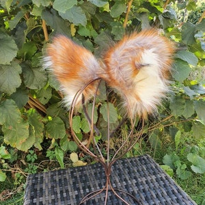 May include: Faux fur fox ears in orange, brown, and white, presented on a copper-coloured wire headpiece. The ears are fluffy and detailed, with a realistic look. The headpiece rests on a dark grey woven surface, with green foliage in the background.