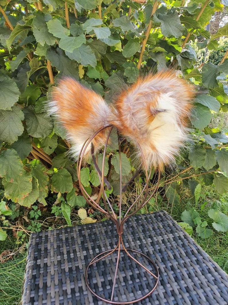 May include: Faux fur fox ears in orange, brown, and white, presented on a copper-coloured wire headpiece. The ears are fluffy and detailed, with a realistic look. The headpiece rests on a dark grey woven surface, with green foliage in the background.