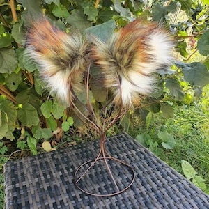 May include: Faux fur fox ears in brown, white, and green, presented on a bronze-coloured metal stand. The ears are fluffy and textured, with a natural colour scheme. The stand features a circular base and a tree-like design.