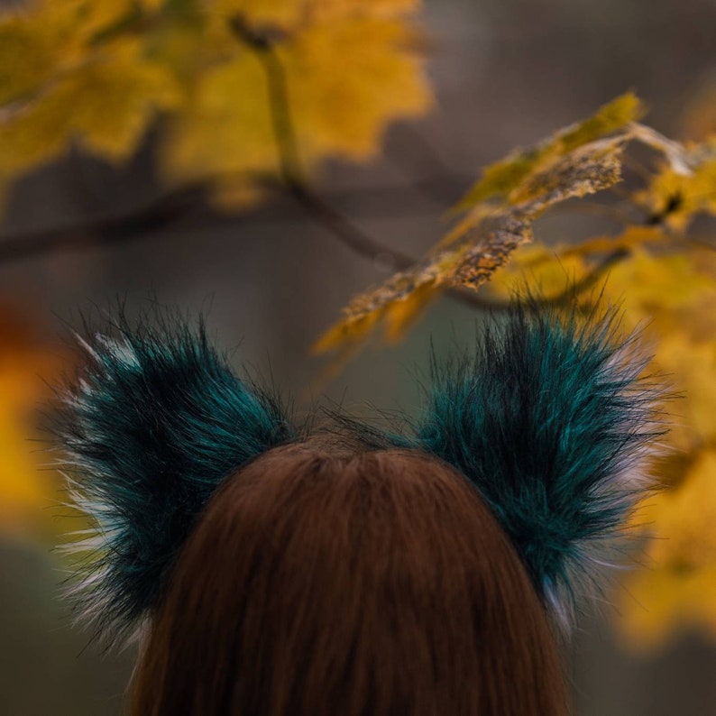 May include: Faux fur ears with teal and black tips, and white accents. The ears are attached to a brown headband. The background shows blurred yellow and green foliage, suggesting an outdoor setting.