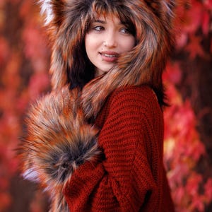 May include: A woman wearing a red jumper and a fluffy fox-like hat with long, brown and black fur. The hat has large, furry ears. The woman is standing in front of a background of red and orange leaves.