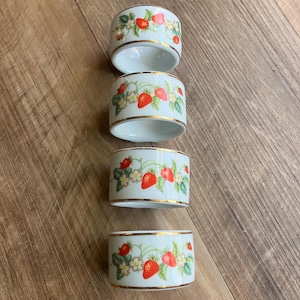 May include: Four white ceramic napkin rings with a strawberry design. Each ring features a repeating pattern of red strawberries, green leaves, and white flowers with gold trim. The napkin rings are stacked on a wooden surface.