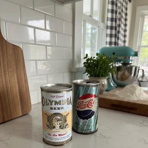 May include: Two vintage beverage cans on a kitchen counter. One is an Olympia Beer can with a gold and white label, and the other is a Pepsi-Cola can with a blue, red, and white design. The cans are 355 ml.