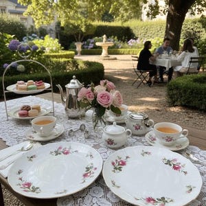 May include: An outdoor tea setting with white plates featuring pink floral designs, teacups, a silver teapot, and a tiered dessert stand with macarons. A vase of pink roses and a sugar bowl complete the elegant arrangement.