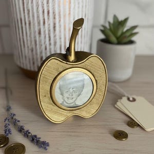 May include: A gold-colored apple-shaped picture frame with a small, round photo inside. The frame has a textured surface and a stem. The photo is a black and white portrait of a person. Decorative buttons and lavender sprigs are nearby.