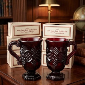 May include: Two ruby red pedestal mugs with ornate detailing and curved handles are displayed on a wooden surface. The mugs are in front of boxes that read "1876 Cape Cod Collection Two Pedestal Mugs."