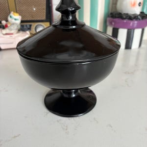 May include: A black glass candy dish with a matching lid featuring a pointed finial handle. The bowl has a smooth, rounded shape and sits on a circular pedestal base. The glass has a glossy, opaque finish that reflects light on its surface.